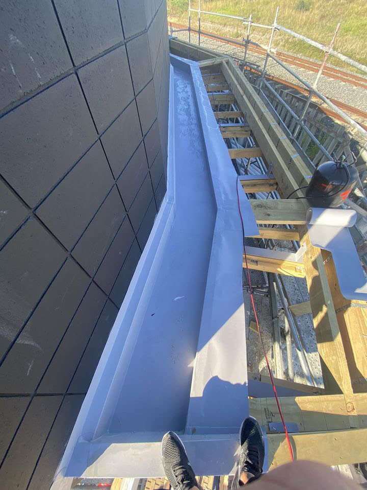 Grey membrane lining a long box gutter beside a dark block wall, seen from the scaffold