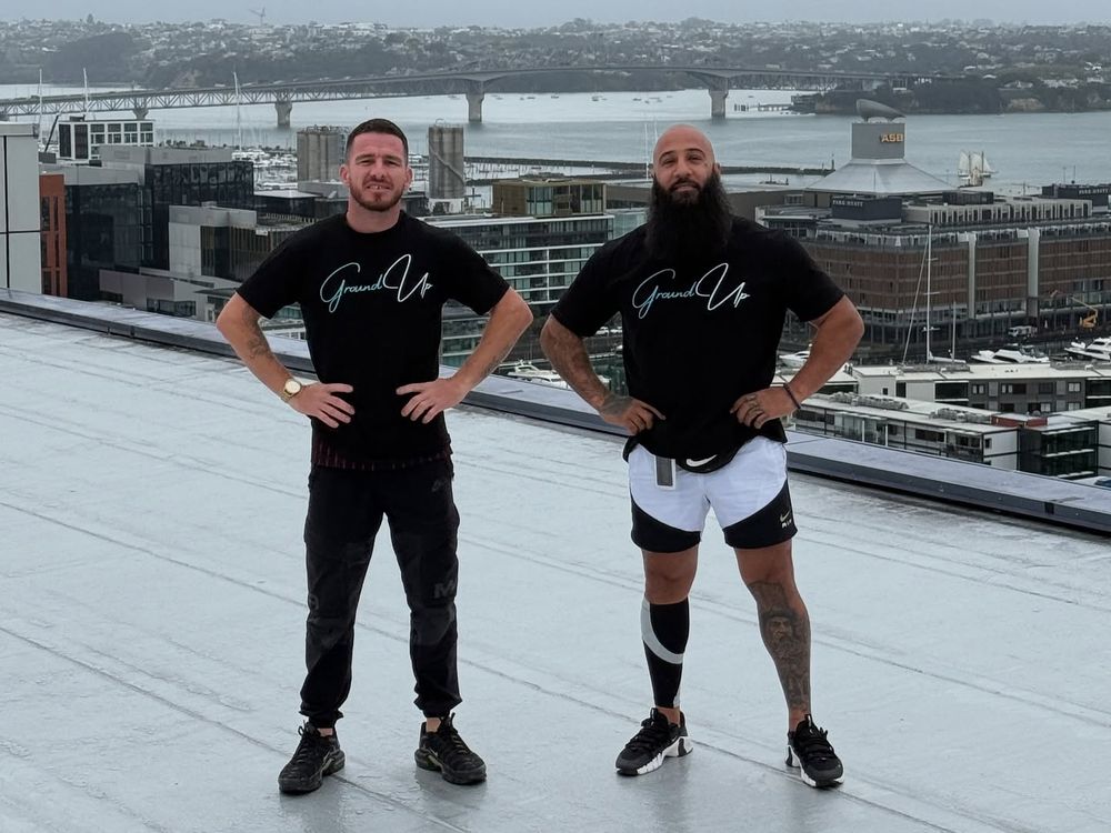 Two of the Ground Up team in branded shirts on a grey membrane roof, with the Auckland waterfront and Harbour Bridge behind